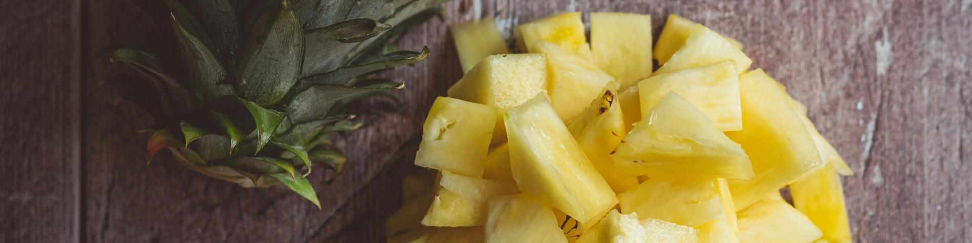 Pieces of pineapple sitting in a bowl on a table, with the crown of a pineapple beside it