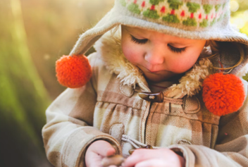 Child wearing beige coat with faux fur trimming and knitted cap with orange pompons