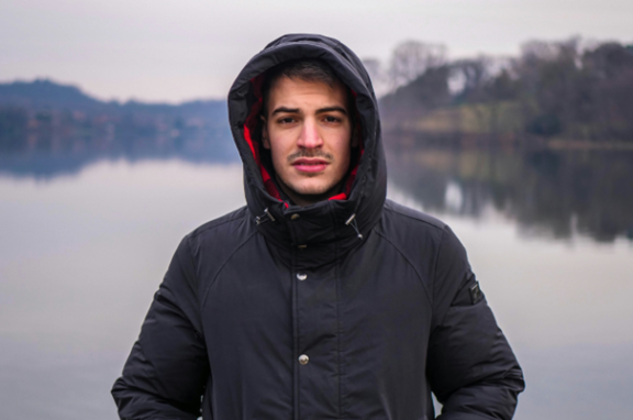 Man wearing a black lacket with the hood up. Behind him is a big lake.